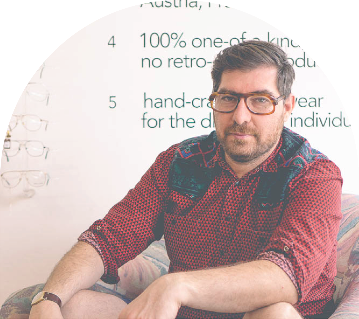 A main in a colorful red shirt wearing eyeglasses sitting in a chair and looking at the camera. 