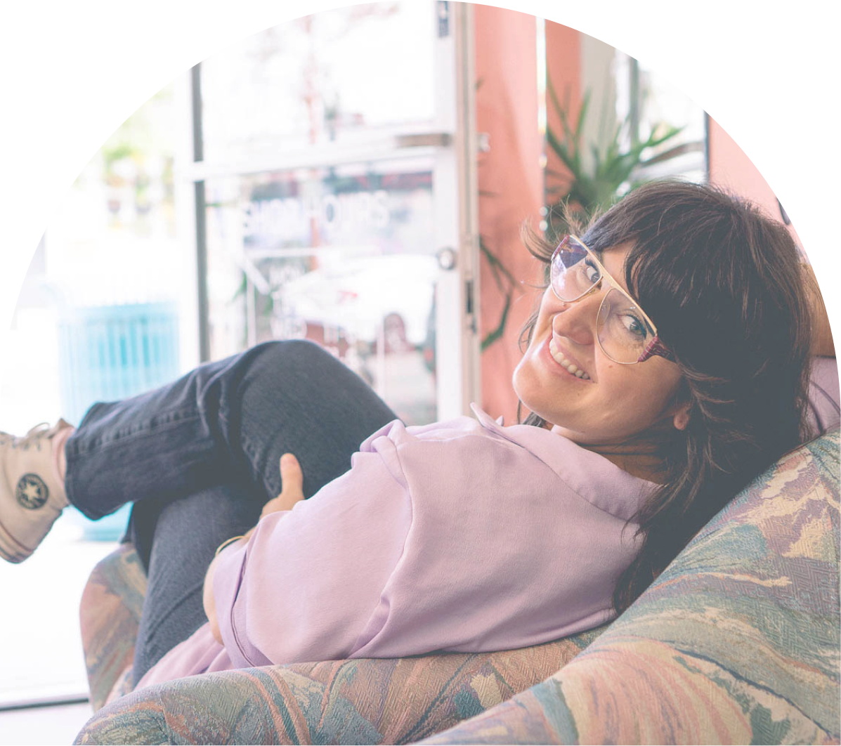 A woman with dark hair wearing a purple shirt and eyeglasses smiling and sitting in a chair. 